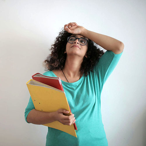 Image of a woman with curly dark hair and greenish-blue shirt holding some binders. She is looking up with her hand on her forehead with a worried look on her face.